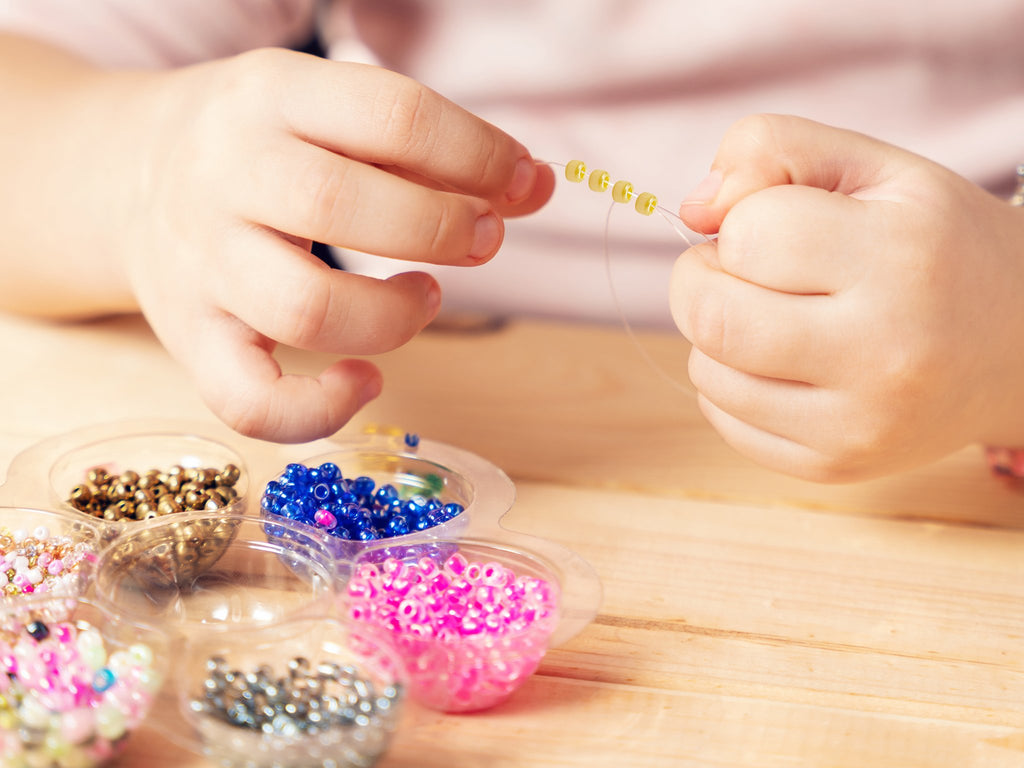 Child's hands stringing yellow seed beads onto clear thread, with containers of colorful beads in foreground.