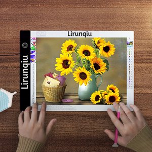 A4 diamond painting LED light pad with sunflowers on a wooden table, hands holding a pen