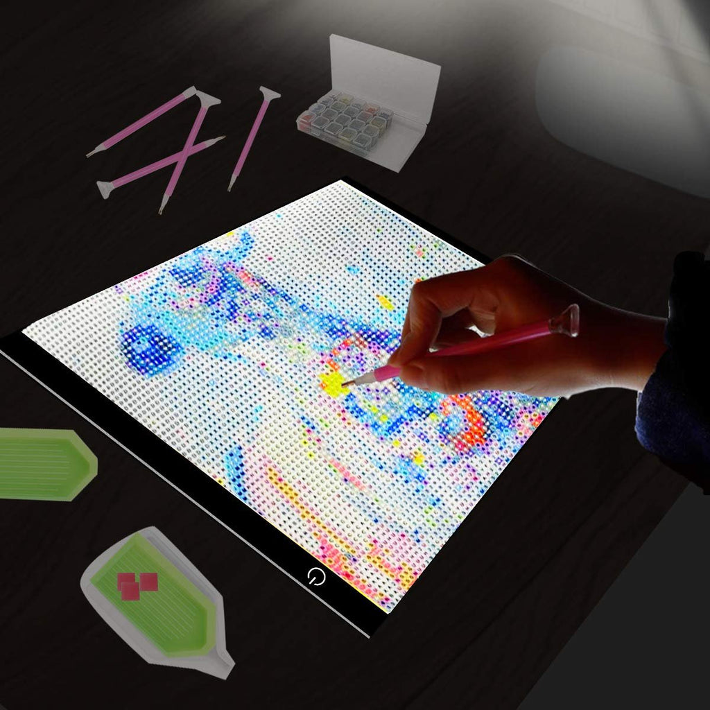 Diamond painting light pad with a hand using a pink stylus to place colorful beads on a partially completed design. Accessories include diamond trays and a storage case.