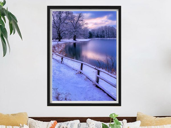 Winter landscape with snow-covered trees and a calm lake reflecting the sky, with a wooden railing along the shore.