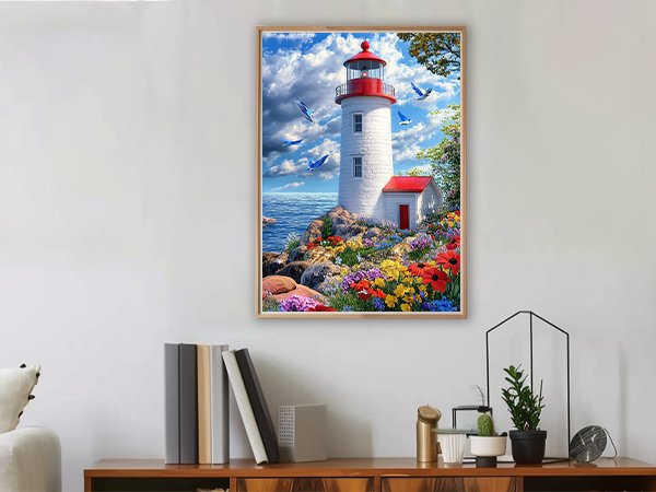 Diamond painting kit of a lighthouse on a rocky shore with colorful flowers and birds flying, framed and displayed on a wall.