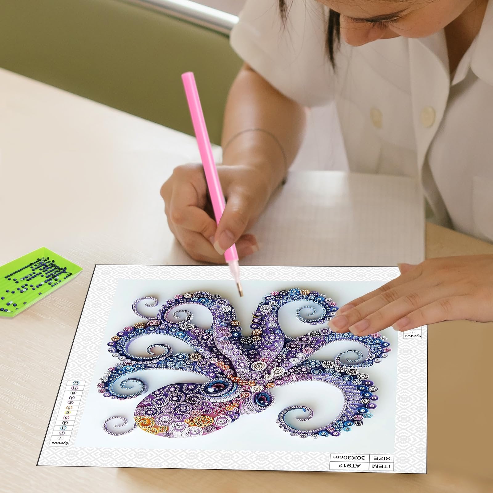 Person completing a diamond art painting of a purple octopus with a pink tool, green diamond sorting tray nearby.