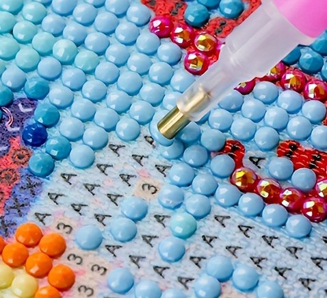 Close-up of a diamond painting kit in progress, showing a tool placing a blue gem onto a numbered canvas.