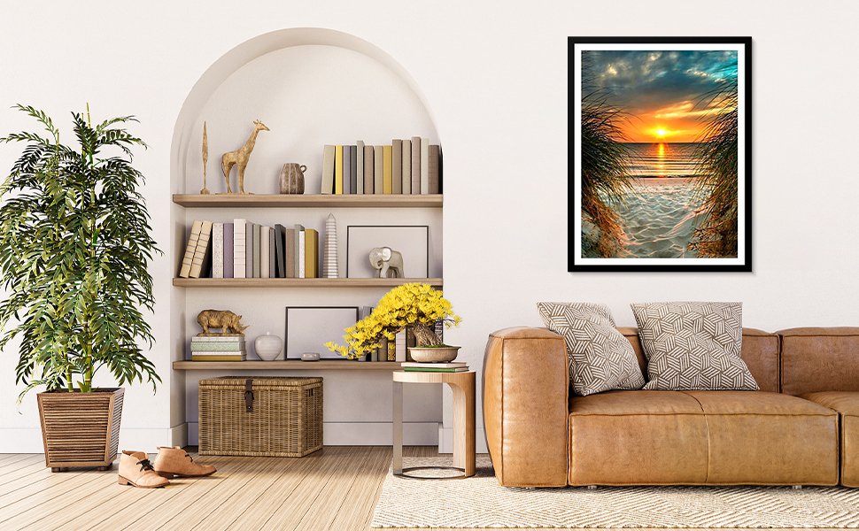 Framed diamond painting of a beach sunset viewed through tall grass, hanging above a leather sofa.