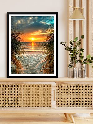 Framed diamond painting of a beach sunset viewed through tall grass, displayed on a wooden cabinet.