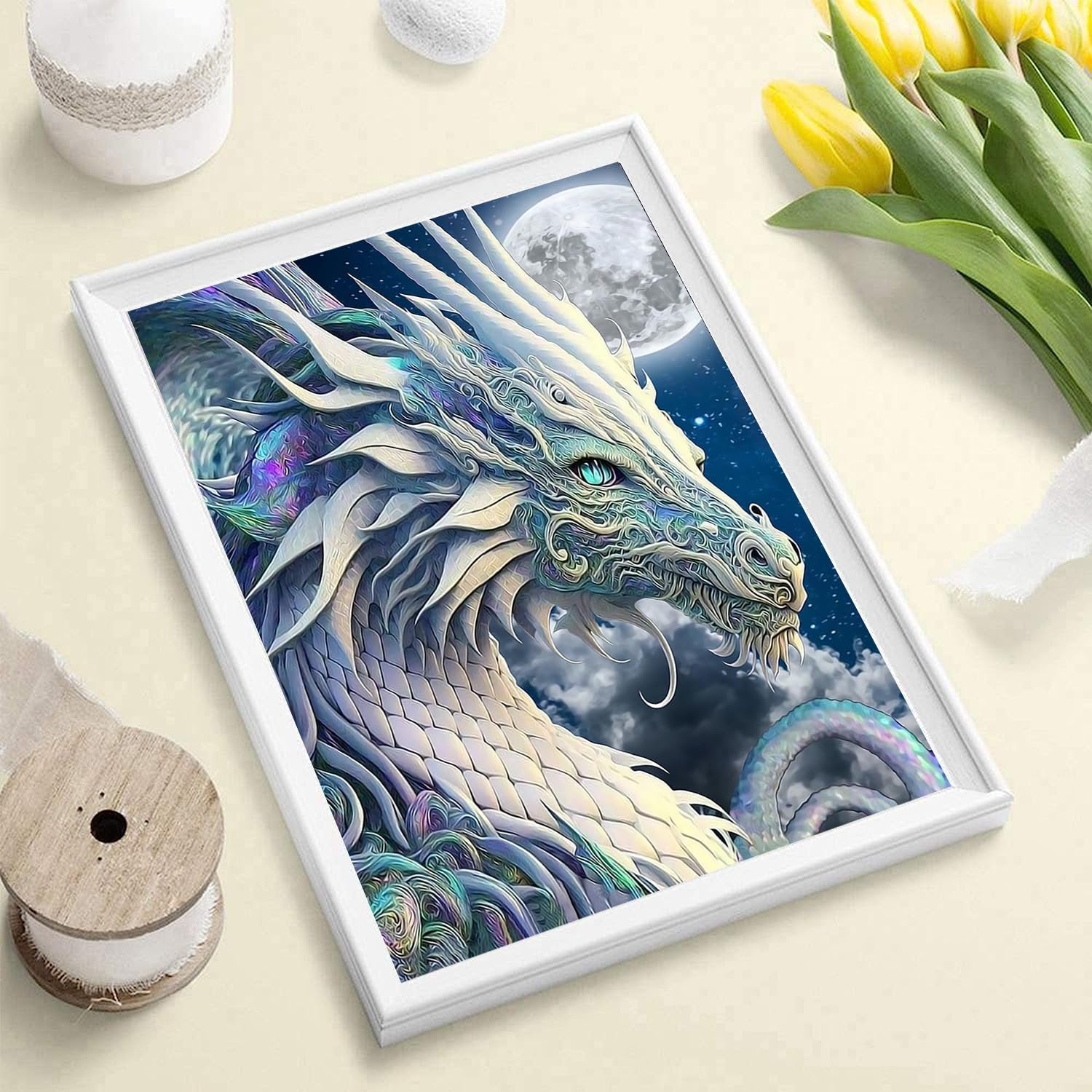 Framed diamond painting kit with a white dragon and moon design, set on a light-colored surface with yellow tulips and a spool of ribbon.