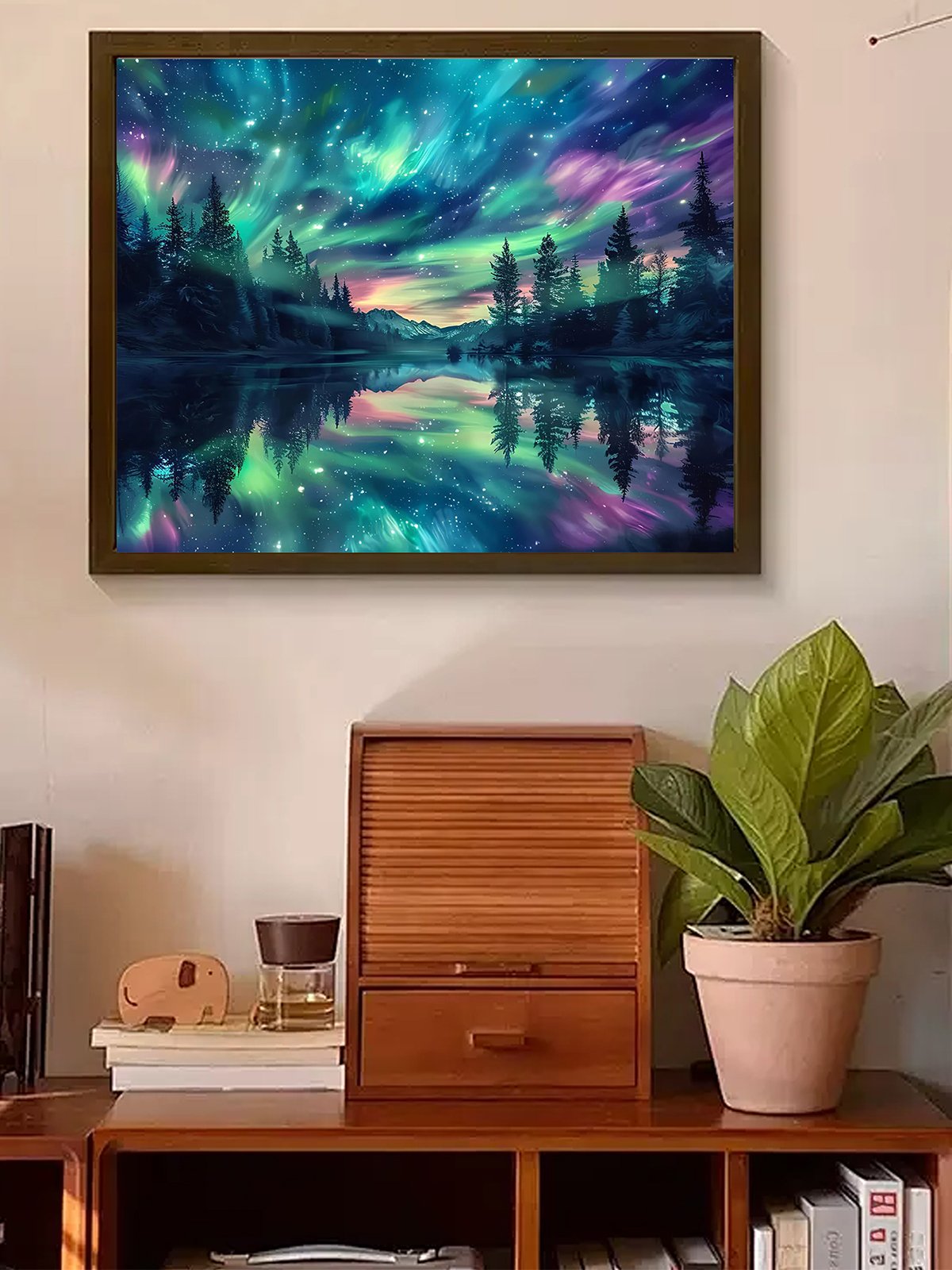 Framed diamond painting of a starry sky aurora borealis over a lake and forest, displayed on a wall above a wooden cabinet.