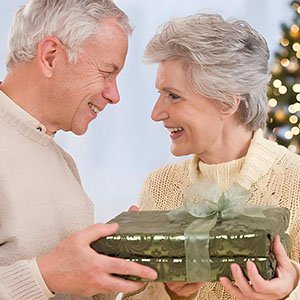 Elderly couple exchanging a wrapped gift box tied with a green ribbon, with a blurred Christmas tree in the background.