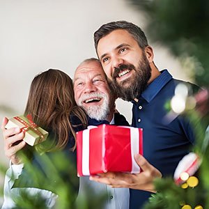 Three people embracing and holding gifts, with a Christmas tree in the foreground.