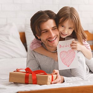 Father and daughter hugging on a bed, daughter holds a handmade card that says 'Daddy' with a heart.