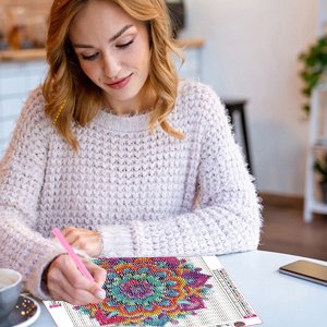Woman with blonde hair wearing a cream sweater, doing a diamond painting kit with a mandala design at a table.