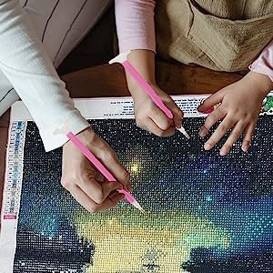 Two people's hands working on a 5D DIY diamond art kit painting of a starry night sky.