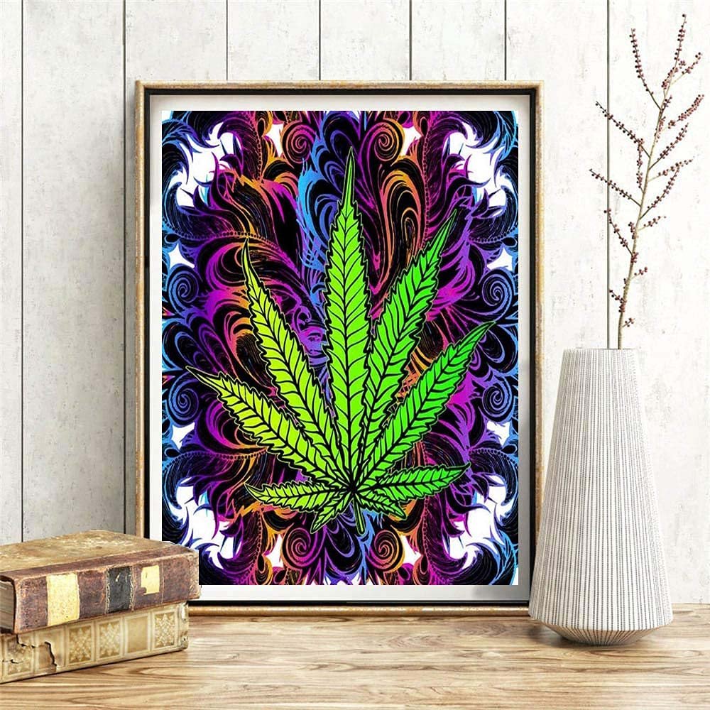 Framed 5D DIY diamond art painting of a green marijuana leaf against a colorful psychedelic background, displayed on a wooden surface with books and a vase.