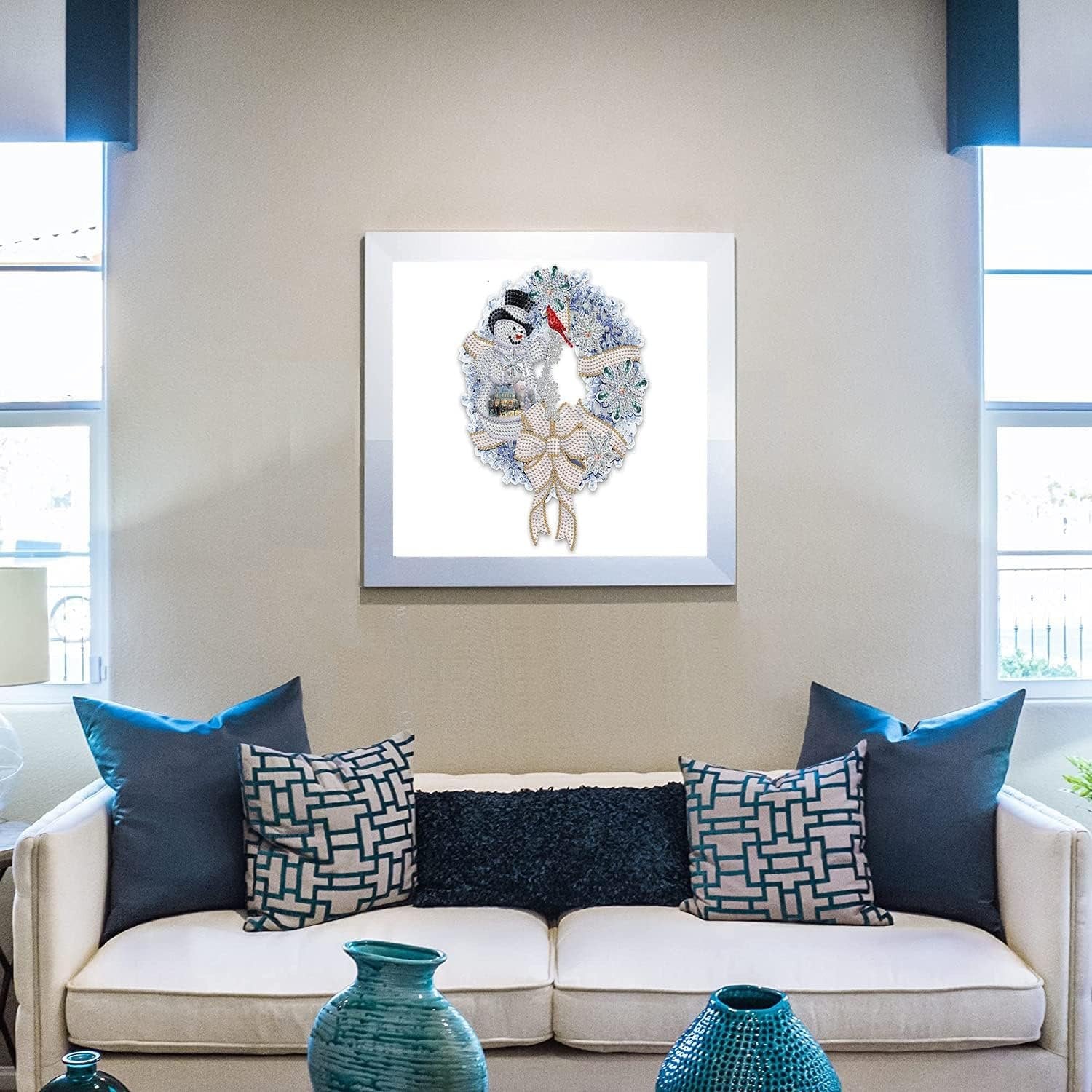 Diamond painting kit of a Christmas wreath with snowmen and a bow, framed and hanging on a wall above a sofa.