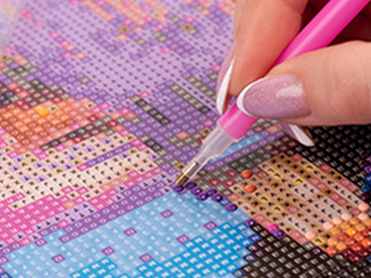 Close-up of a hand using a pink diamond art pen to place a blue diamond onto a canvas with a skullhead design.