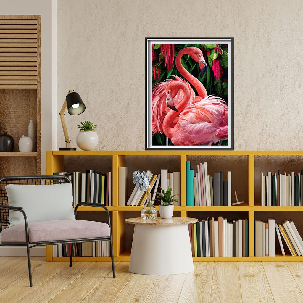 Two pink flamingos with lush green foliage and red flowers, framed artwork on a bookshelf.