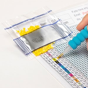 Diamond painting kit showing a hand holding a tool to pick up yellow beads from a small bag onto a numbered grid.