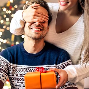 Woman surprising man with an orange gift box, with a Christmas tree blurred in the background.