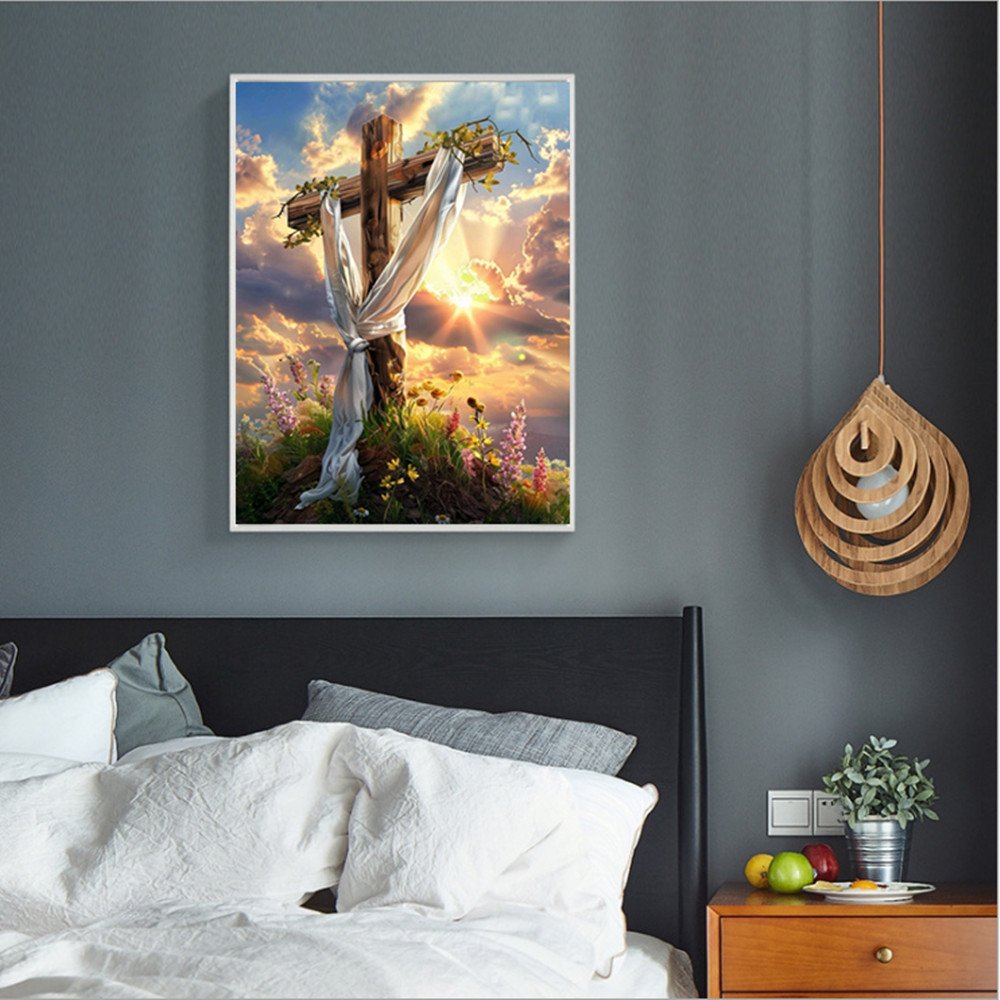 Diamond painting of a wooden cross with draped white cloth and flowers at sunrise, displayed in a bedroom.