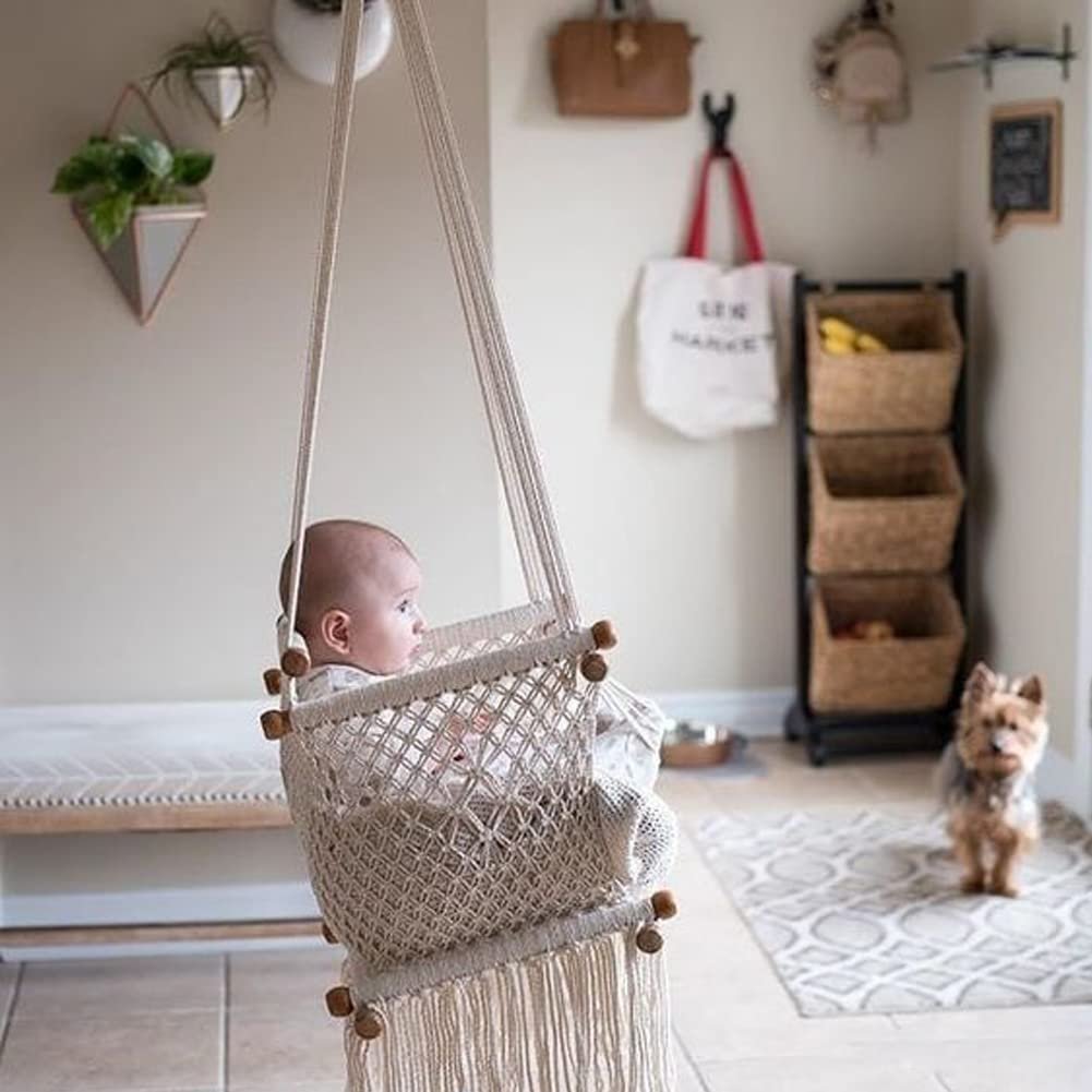 Baby sitting in a beige macrame swing with fringe, a dog stands nearby in a room with baskets and plants.