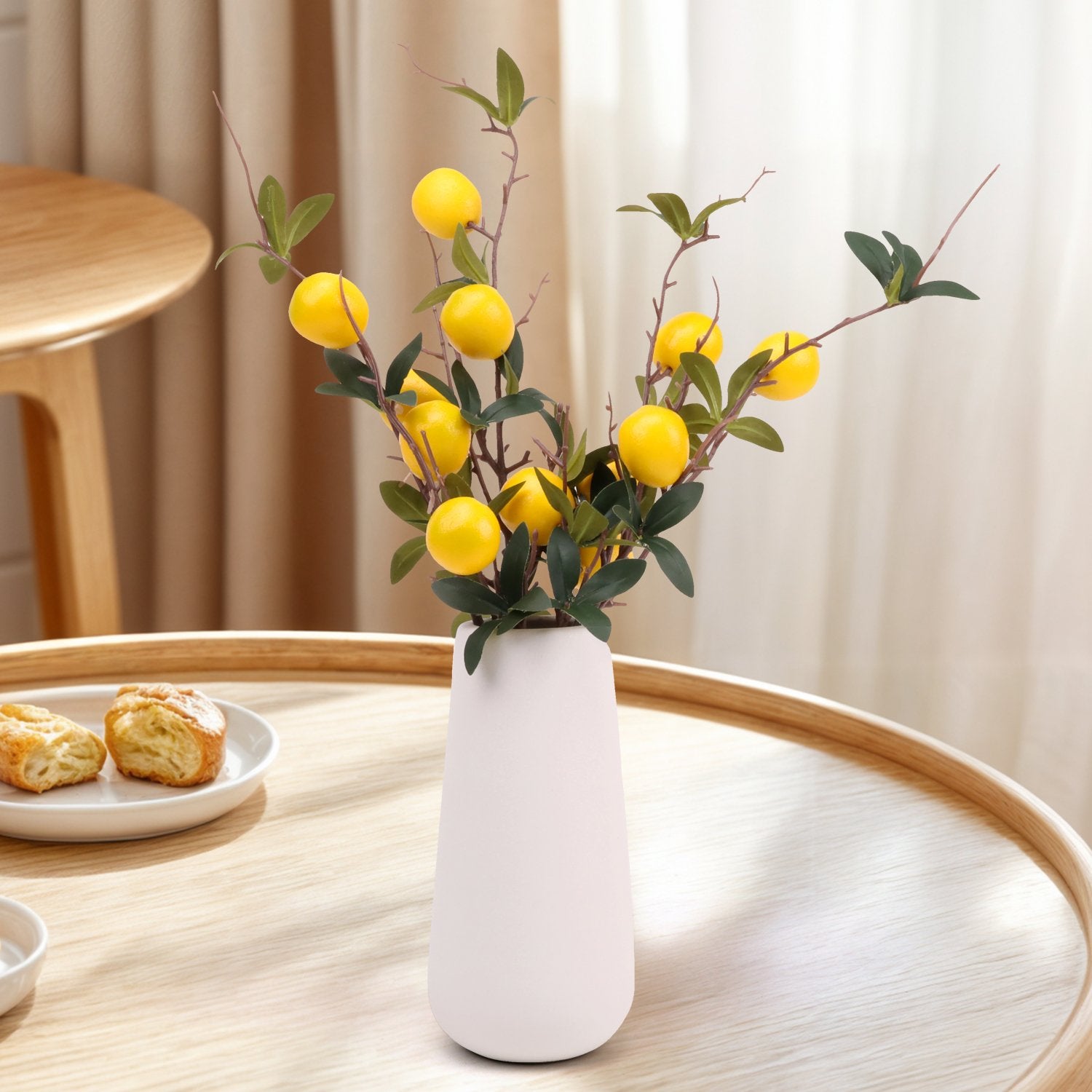 Artificial pomegranate branches with yellow fruit and green leaves in a white vase on a wooden table.