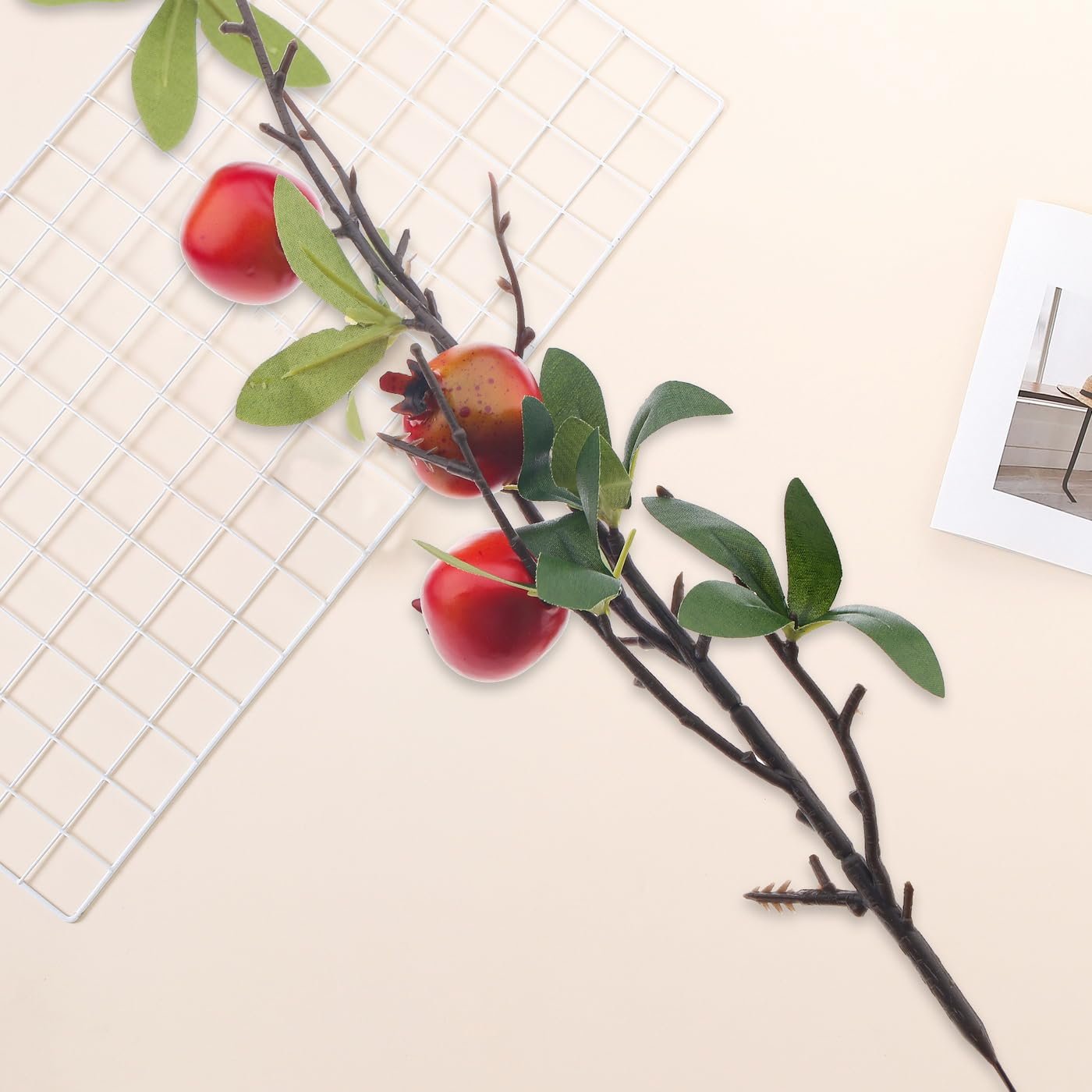 Artificial pomegranate branch with red fruit and green silk leaves on a light background with a white grid and a photo frame.