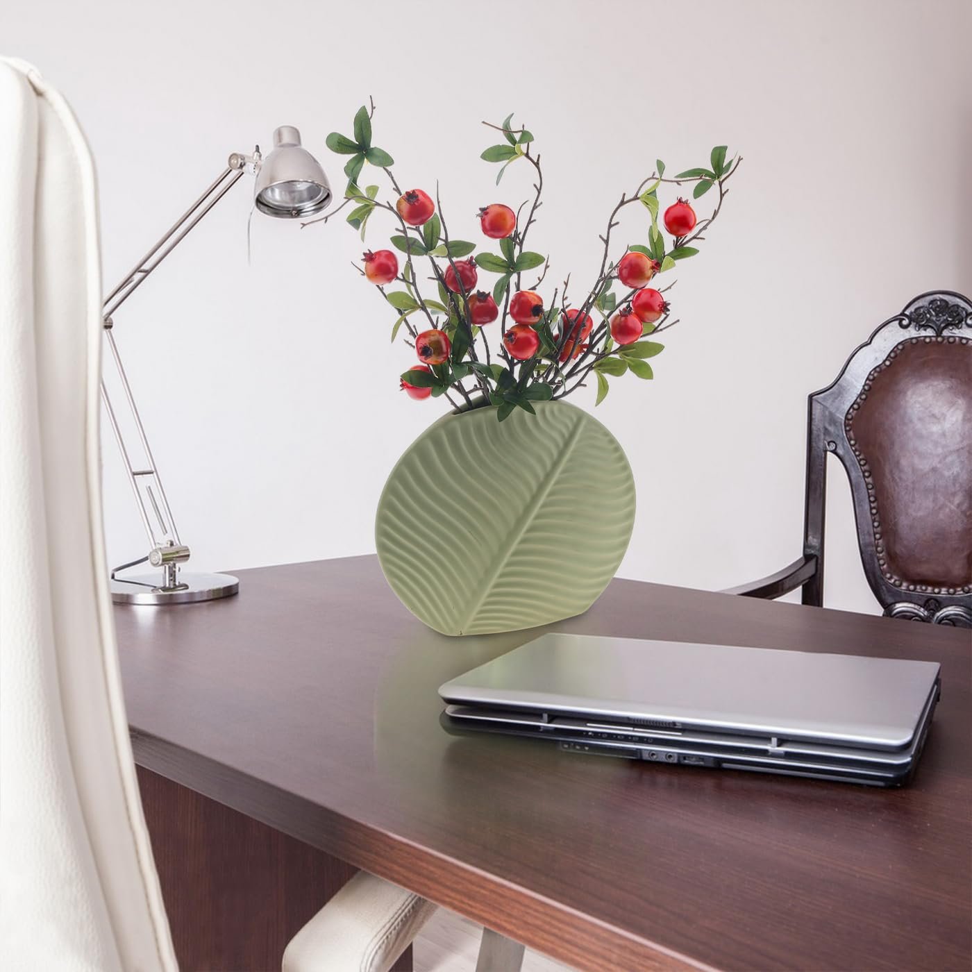 Artificial pomegranate branches with red berries and green leaves in a light green vase on a desk with a laptop and lamp.