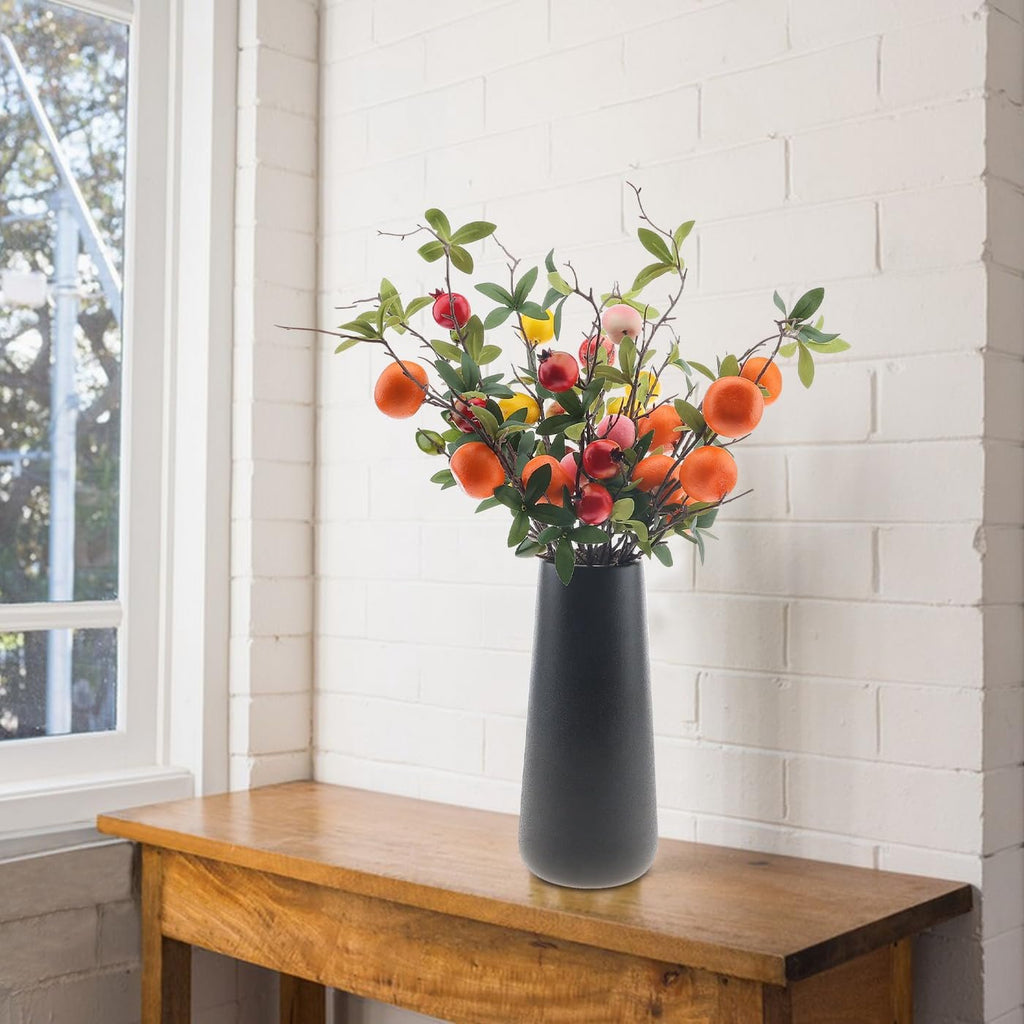 Artificial pomegranate branches with silk leaves and foam fruits in a dark vase on a wooden table by a window.