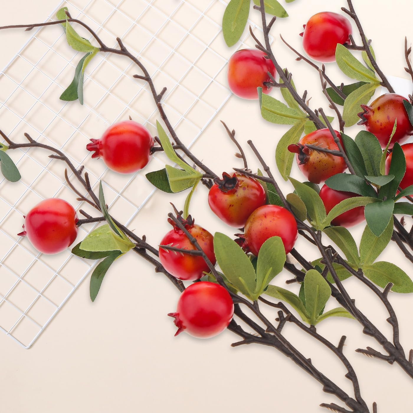 Artificial pomegranate branches with red fruit and green leaves on a cream background with a white grid.