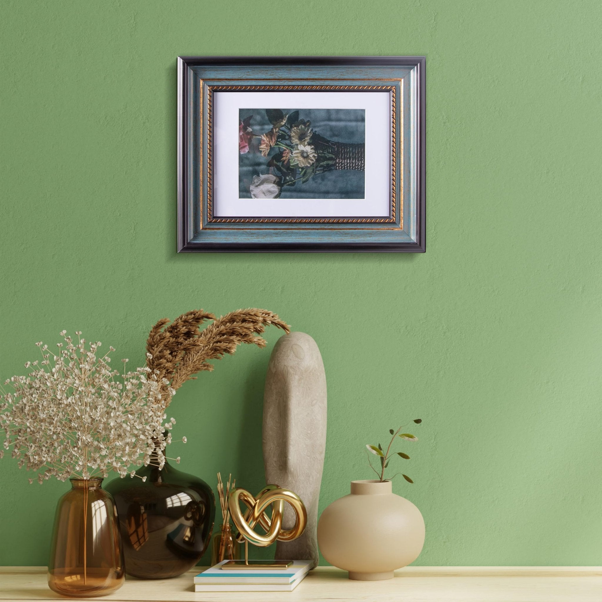 Peacock blue picture frame with gold trim displaying floral art, hung on a green wall above decorative vases and sculptures.