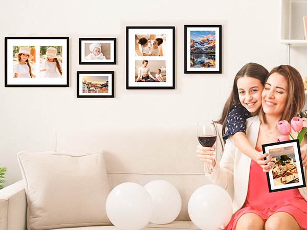 Mother and daughter embracing, holding a picture frame with a scenic view, surrounded by framed photos on a wall.