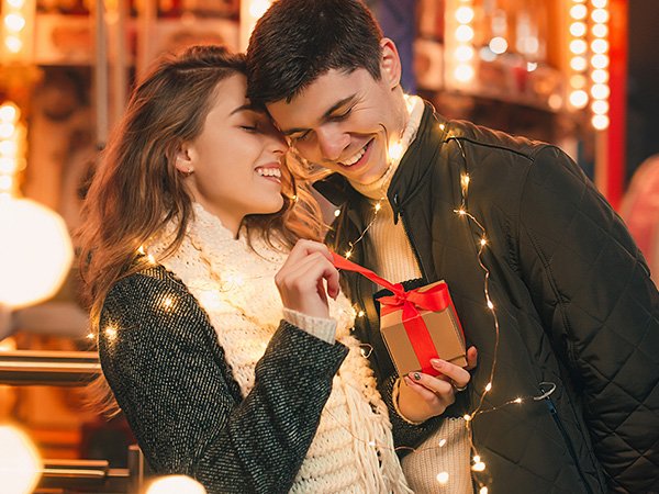 Couple exchanging a gift box with red ribbon, surrounded by fairy lights