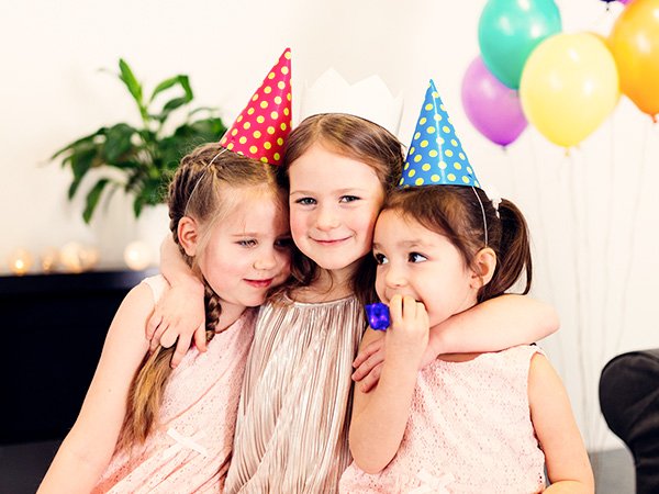 Three young girls in party hats hugging, with balloons in the background.