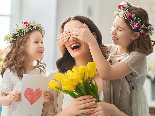 Mother surprised by daughters holding flowers and a handmade card with a heart