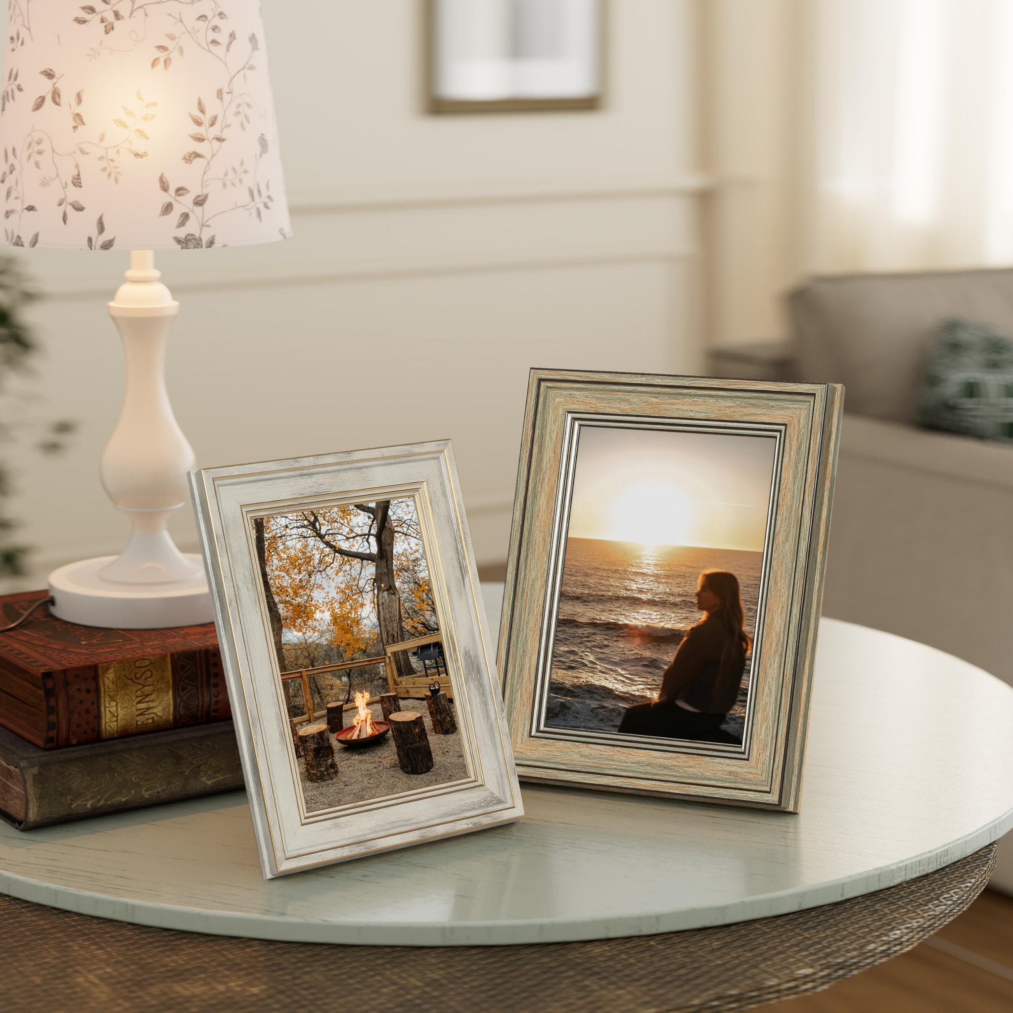 Two rustic farmhouse picture frames on a table with a lamp and books, one frame shows a campfire scene, the other a sunset over the ocean.