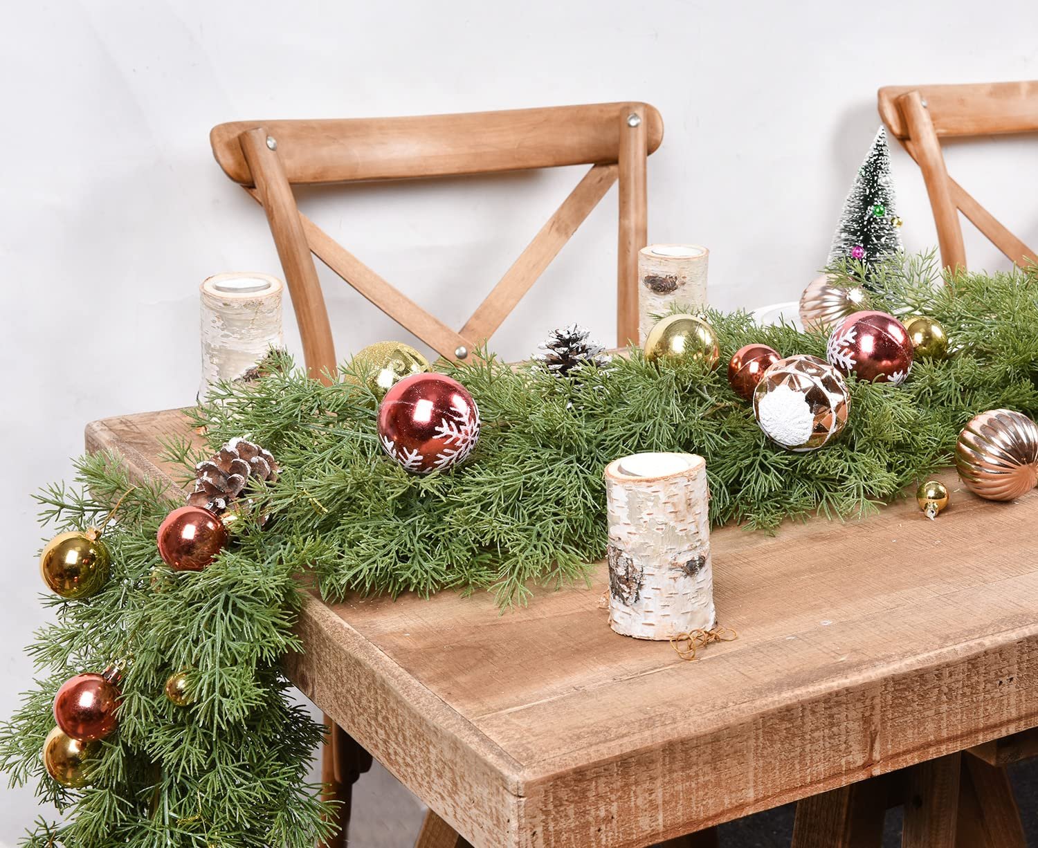 Artificial pine Christmas garland with red and gold ornaments and pine cones draped over a wooden table between two chairs.