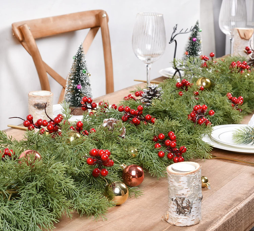 Artificial pine Christmas garland with red berries and gold ornaments on a wooden table setting.