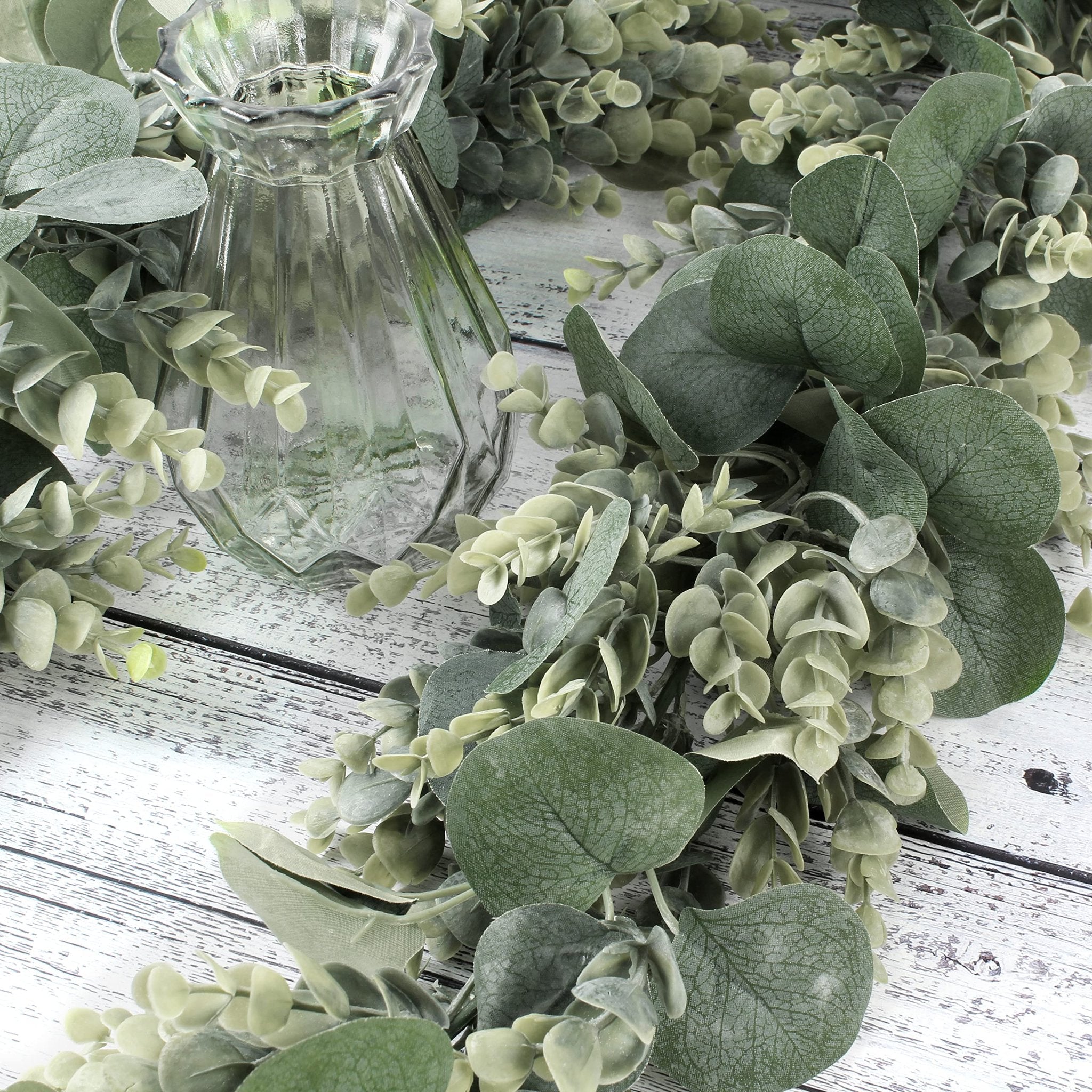 Artificial eucalyptus and lambs ear garland draped around a clear glass vase on a white wooden surface