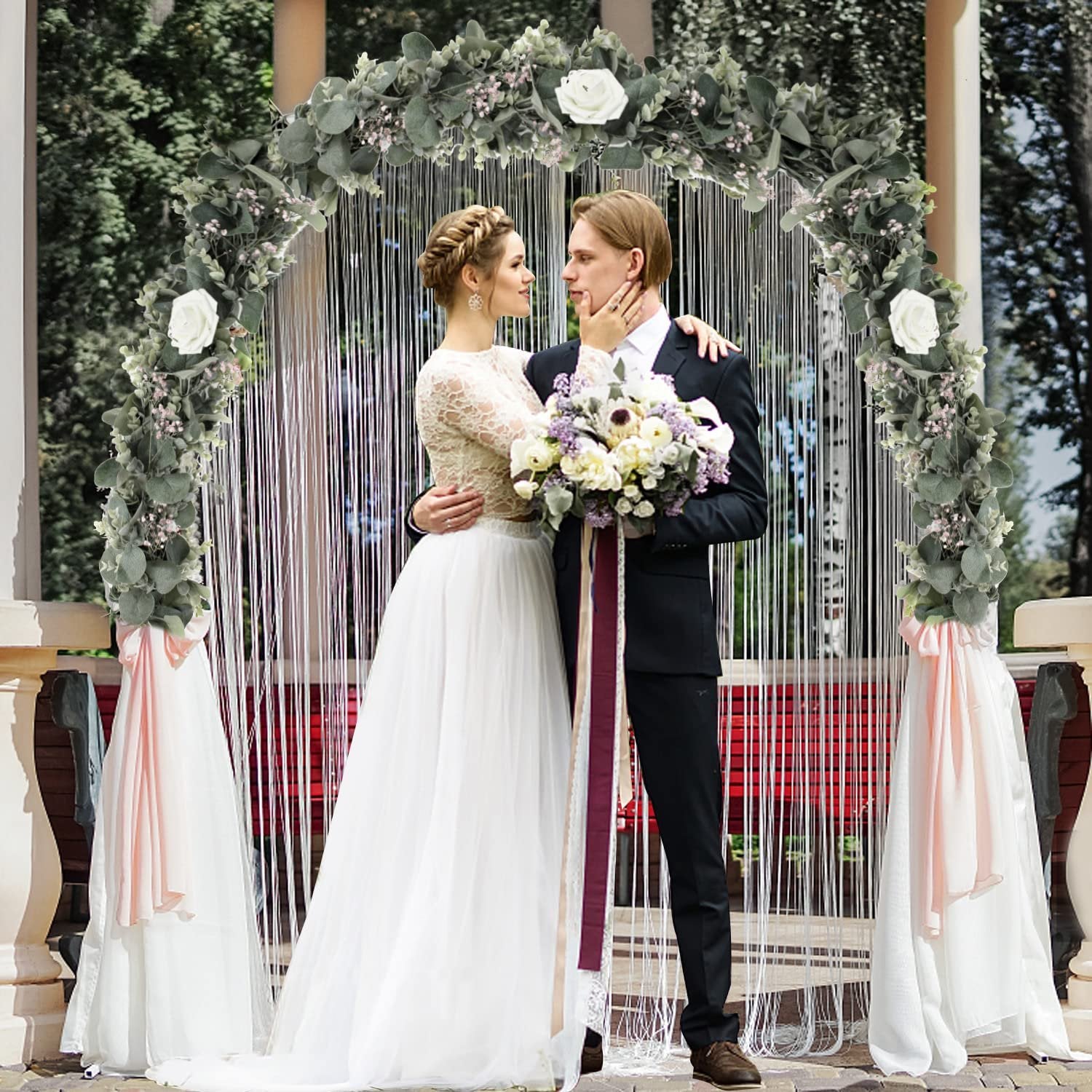 Bride and groom embracing under a wedding arch decorated with faux eucalyptus, lamb's ear, and magnolia leaves.