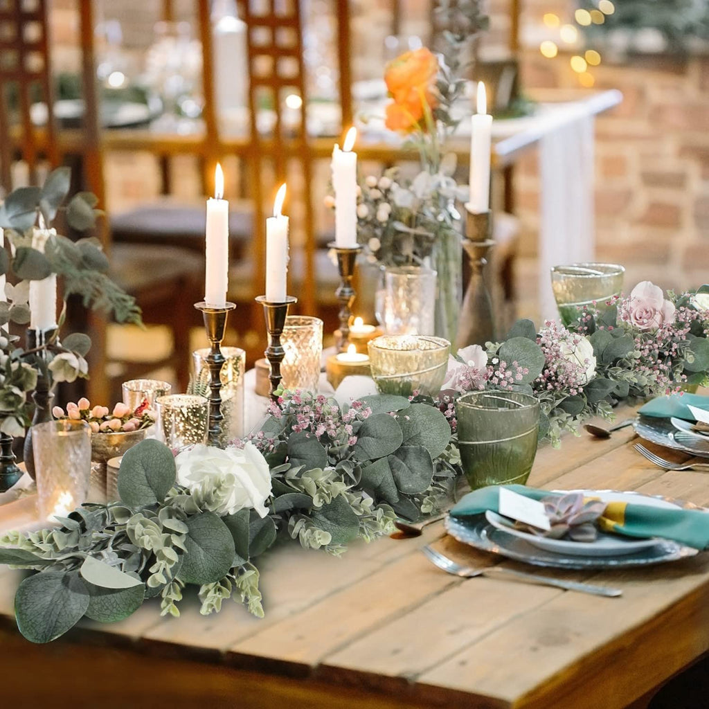 Faux eucalyptus garland with white roses and pink baby's breath draped across a wooden table set for a meal.