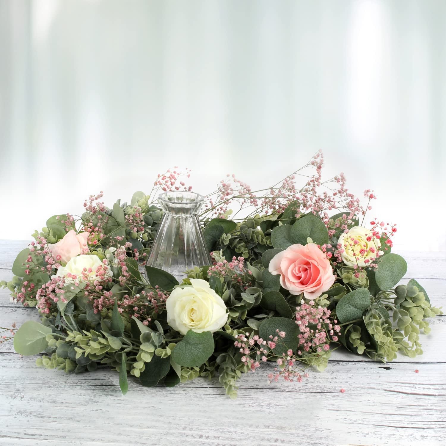 Faux eucalyptus garland with pink and white roses and baby's breath arranged around a glass vase on a white wooden surface.