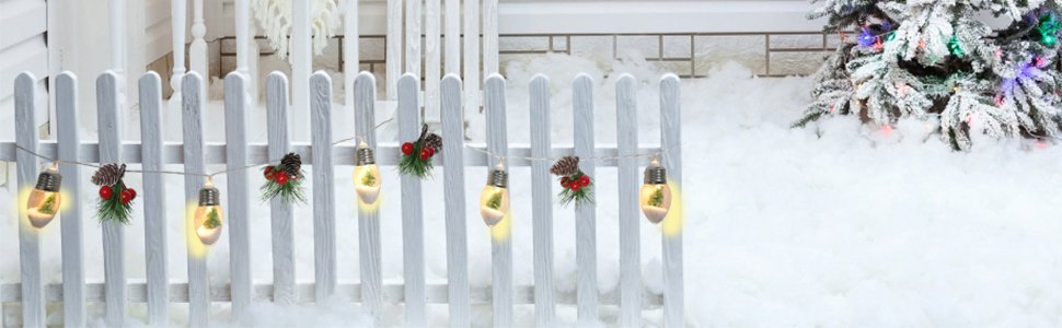 Christmas pine garland with lights and pinecone ornaments draped on a white picket fence with snow and a decorated tree in the background.
