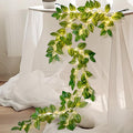 Green vine garland with 96 LED lights draped over a white draped surface, showcasing its naturalistic design and warm illumination.