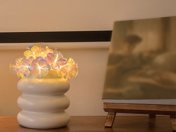 Fiber optic flower string lights in a white vase on a wooden surface, with a blurred canvas on an easel to the right.