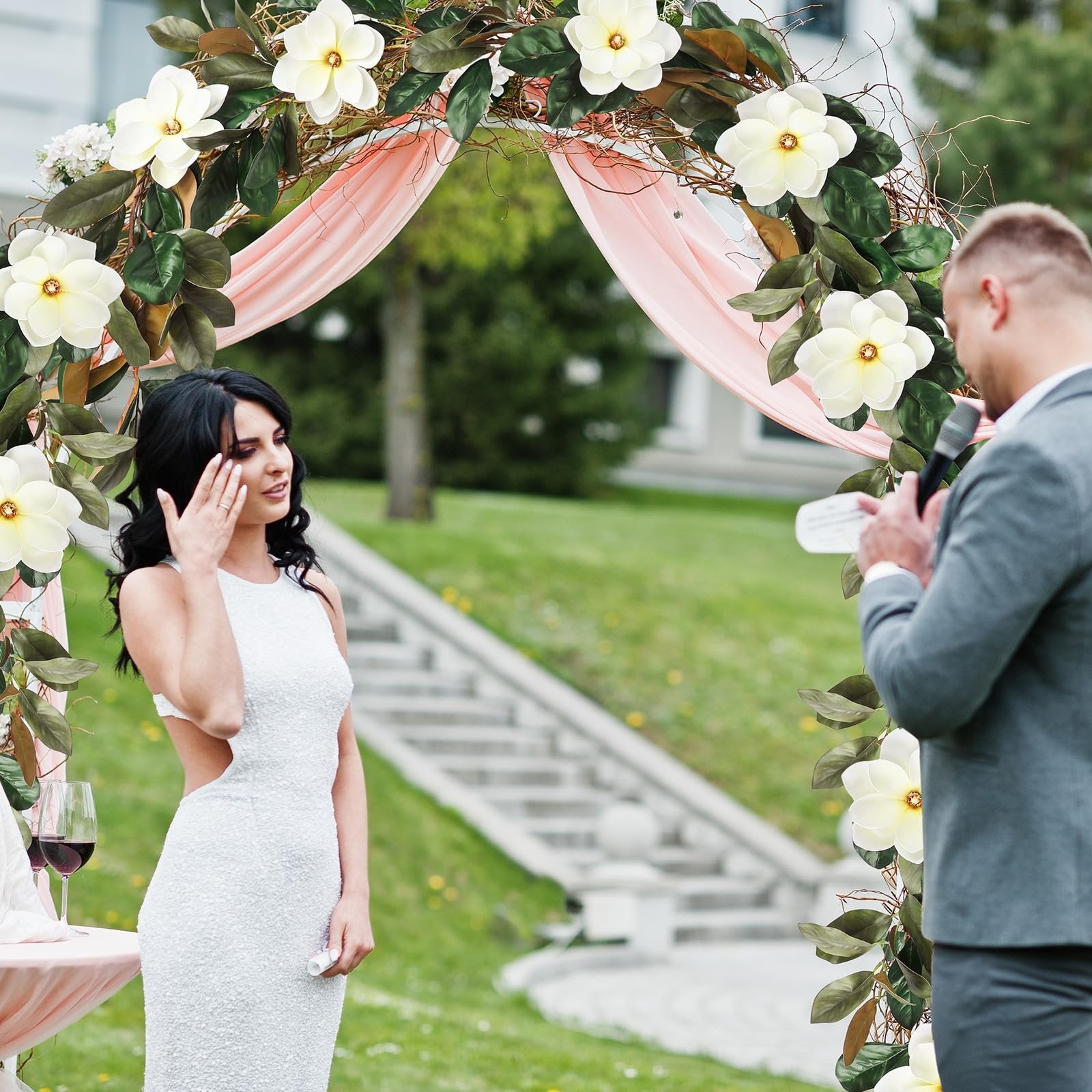 Wedding ceremony with artificial magnolia garland arch, bride in white dress, groom speaking into microphone.