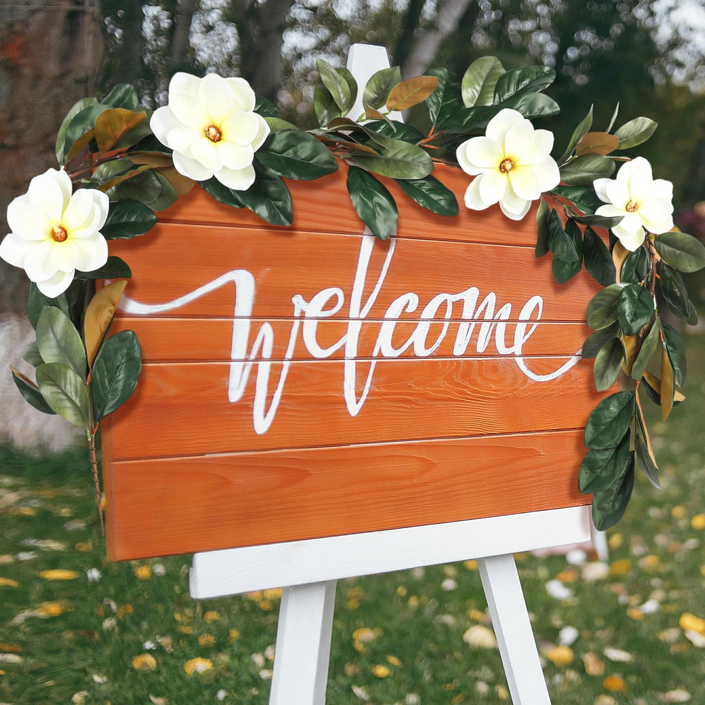 Orange wooden welcome sign decorated with artificial magnolia garland and white flowers on a white easel outdoors.