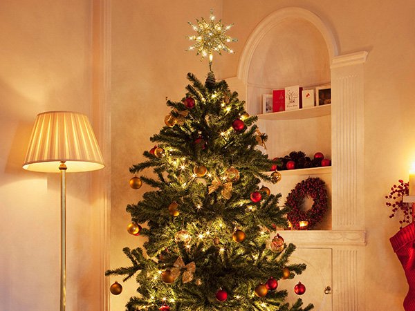 Lit Christmas tree with a starburst tree topper and ornaments, next to a floor lamp and decorative shelf.