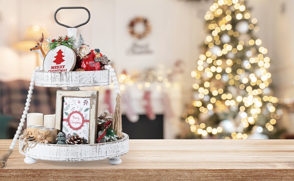 Two-tiered distressed white wooden tray with Christmas decorations, including a reindeer, pinecones, candles, and a framed Merry Christmas card, set against a blurred background of a Christmas tree and fireplace.
