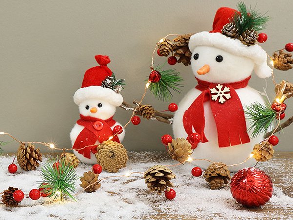Two plush snowmen wearing red hats and scarves, decorated with pinecones and berries, with a string of warm white LED lights.