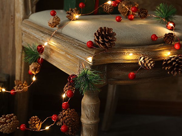 Christmas garland with pinecones, red berries, bells, and warm white LED lights draped over a wooden chair.